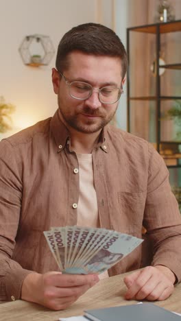 businessman freelancer holding fan of hryvnias cash showing thumbs up sitting at home office table