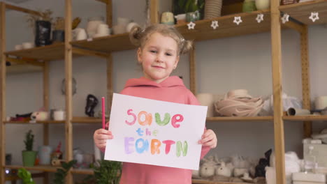 blonde little girl in craft workshop holding a cardboard sign with save the earth phrase 1