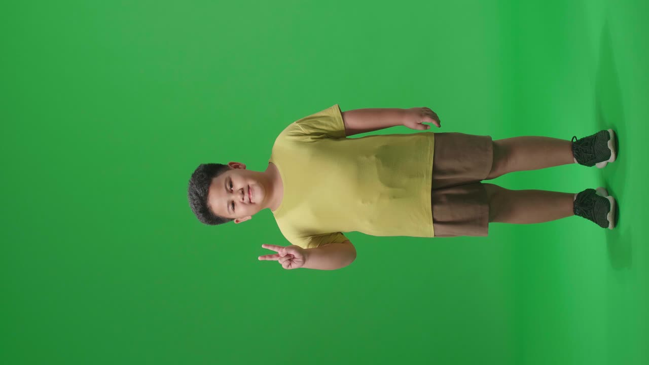 Full Body Of A Smiling Asian Little Boy Showing Gesture Peace While Standing On Green Screen In The Studio