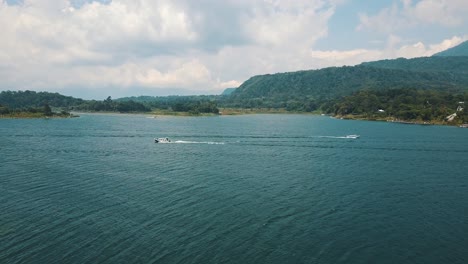 drone aerial flying over trees towards lake atitlan with traveling boats in guatemala, central america