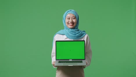 asian muslim woman smiling and showing green screen laptop to camera while standing in the green screen background studio