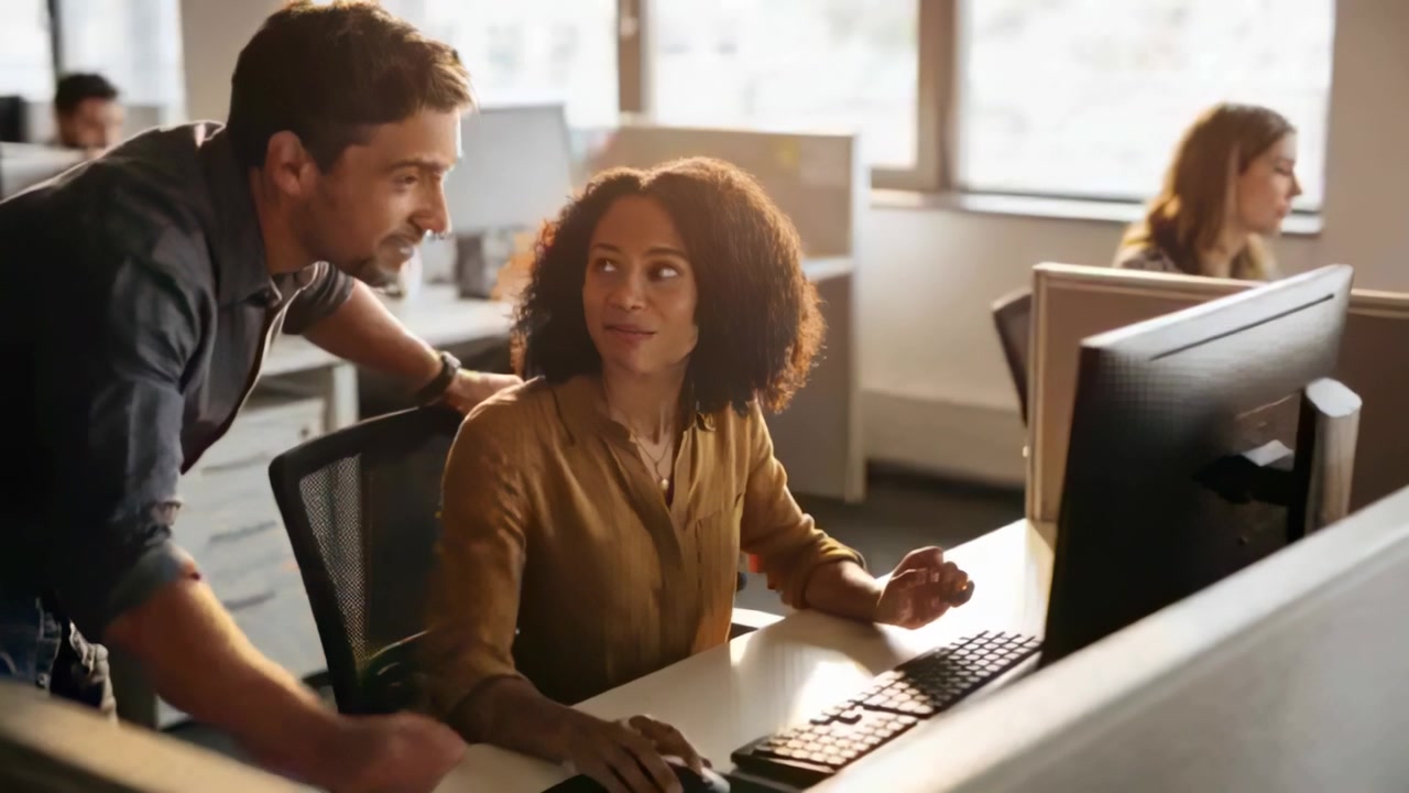 Typing woman brown top pausing at desk, male coworker leaning in gesturing at monitor, helping