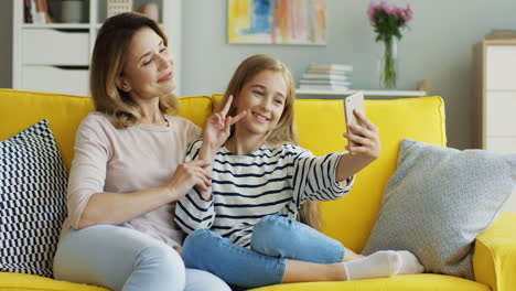 close up view of mother and cute girl taking a selfie with smartphone while they showing ok gesture with her hands sitting on sofa in living room