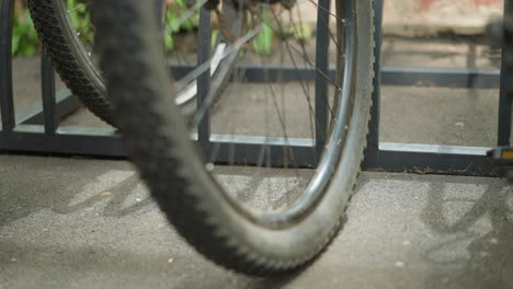 primer plano de una rueda de bicicleta estacionada con seguridad en un soporte de bicicleta de metal, enfatizando los radios y el marco, con detalles borrosos de las bicicletas circundantes y la vegetación en el fondo