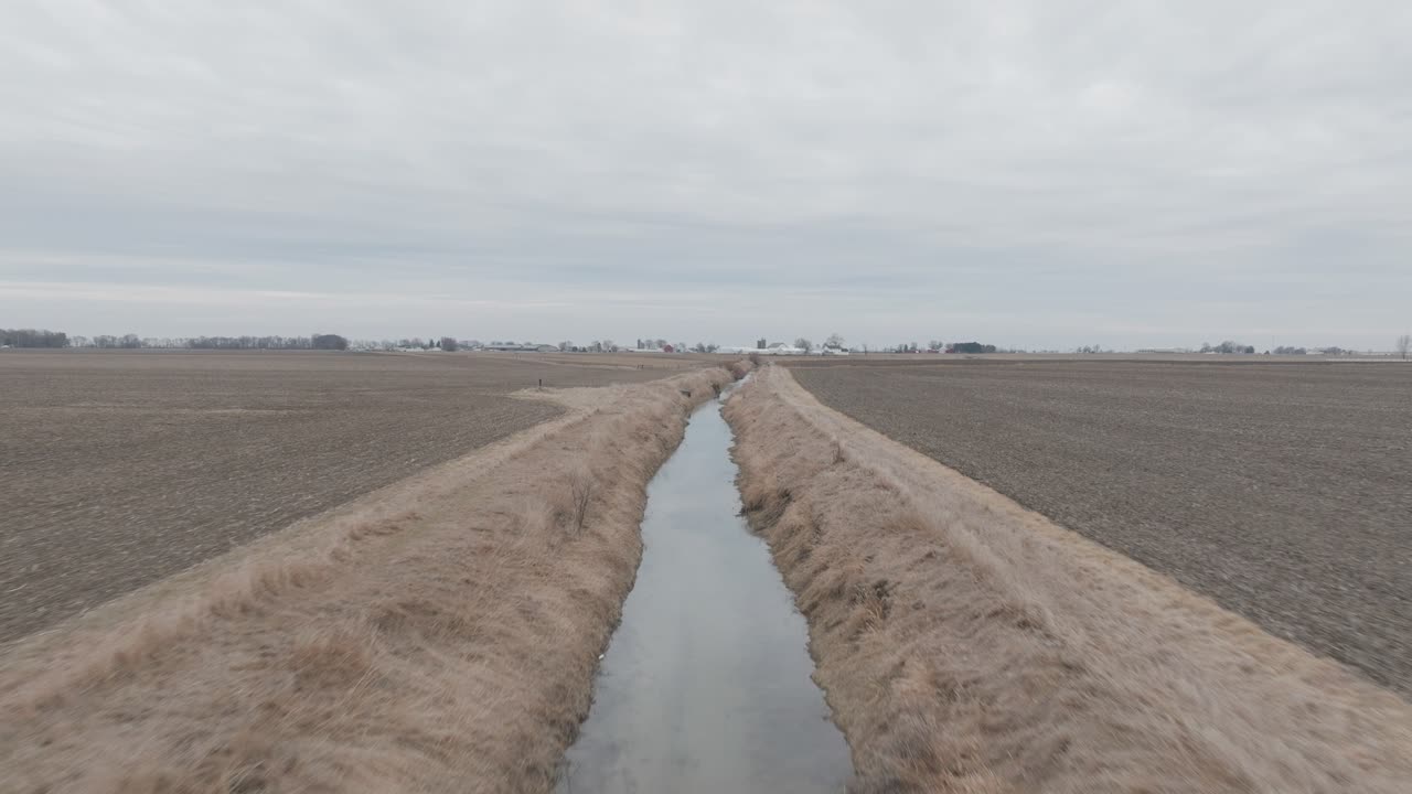 Aerial shot of long stretched ditch filled with water cutting through dry, grassy field.