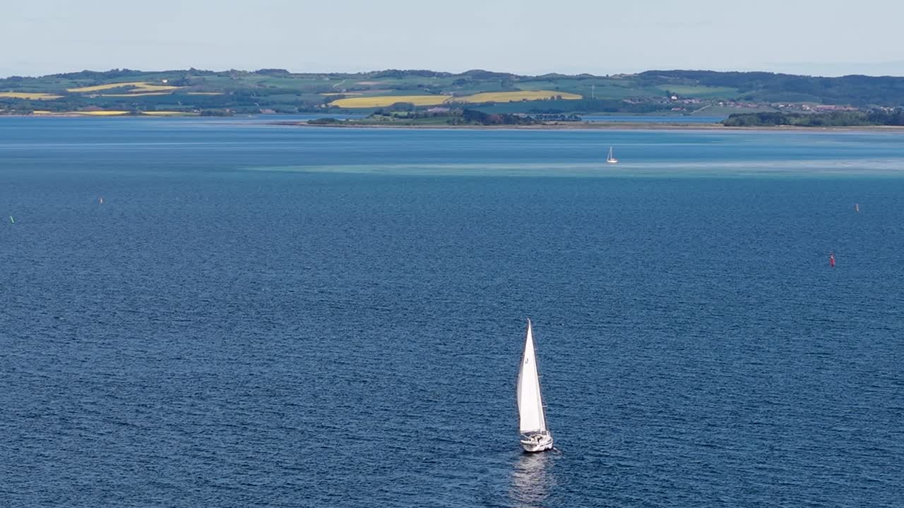 Aerial drone view of a lone sailboat gliding across deep blue waters with distant green shoreline in the background