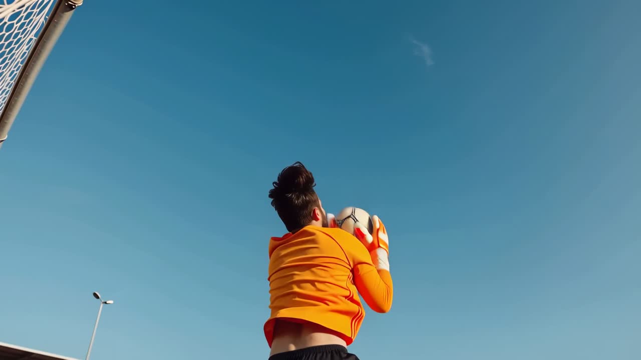 Portiere in estensione con maglia arancione che afferra il pallone da partita in arrivo in porta, preparandolo per un passaggio.