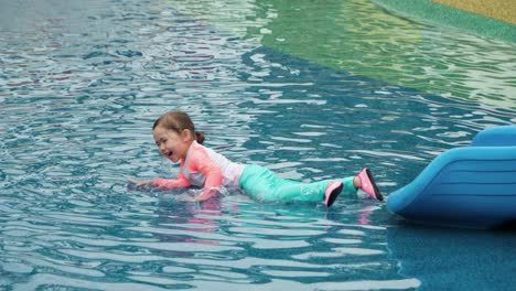 happy little toddler in swimsuit refreshing lying in shallow water, gets up, playful girl jumps and smiles at water park playground - slow motion