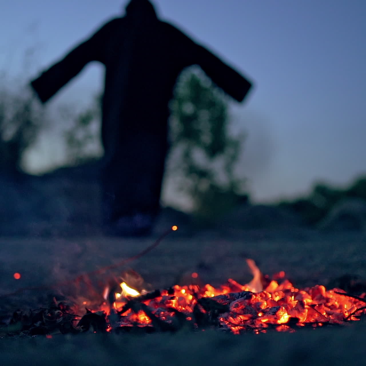 Embers of burnt-out fire and black witch outdoors. Dark death near smoldered logs in the evening. Mysterious figure on the natural background.
