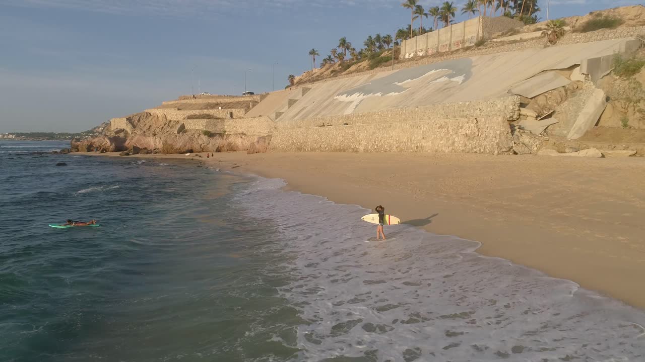 серфер приближается к воде на пляже с белой доской для серфинга на восходе солнца, воздушная точка интереса