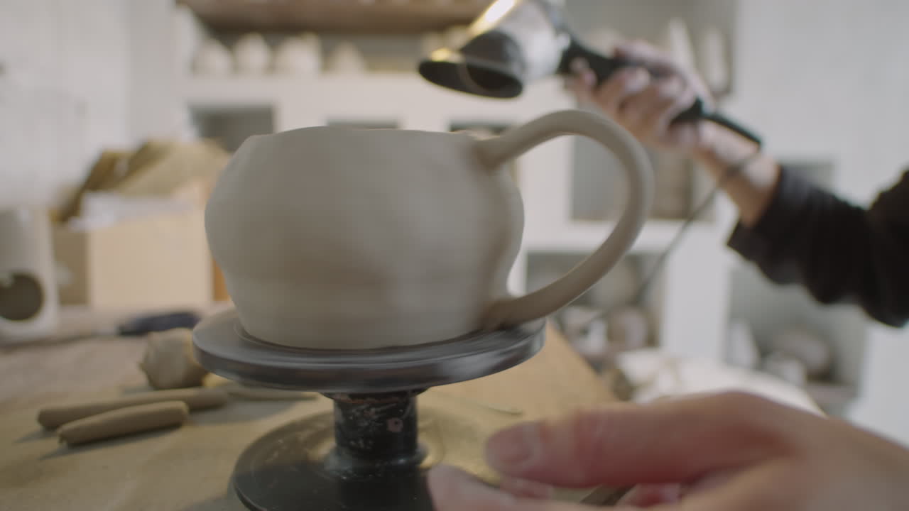 Hands of Artisan Drying Ceramic Mug on Pottery Wheel with Hairdryer