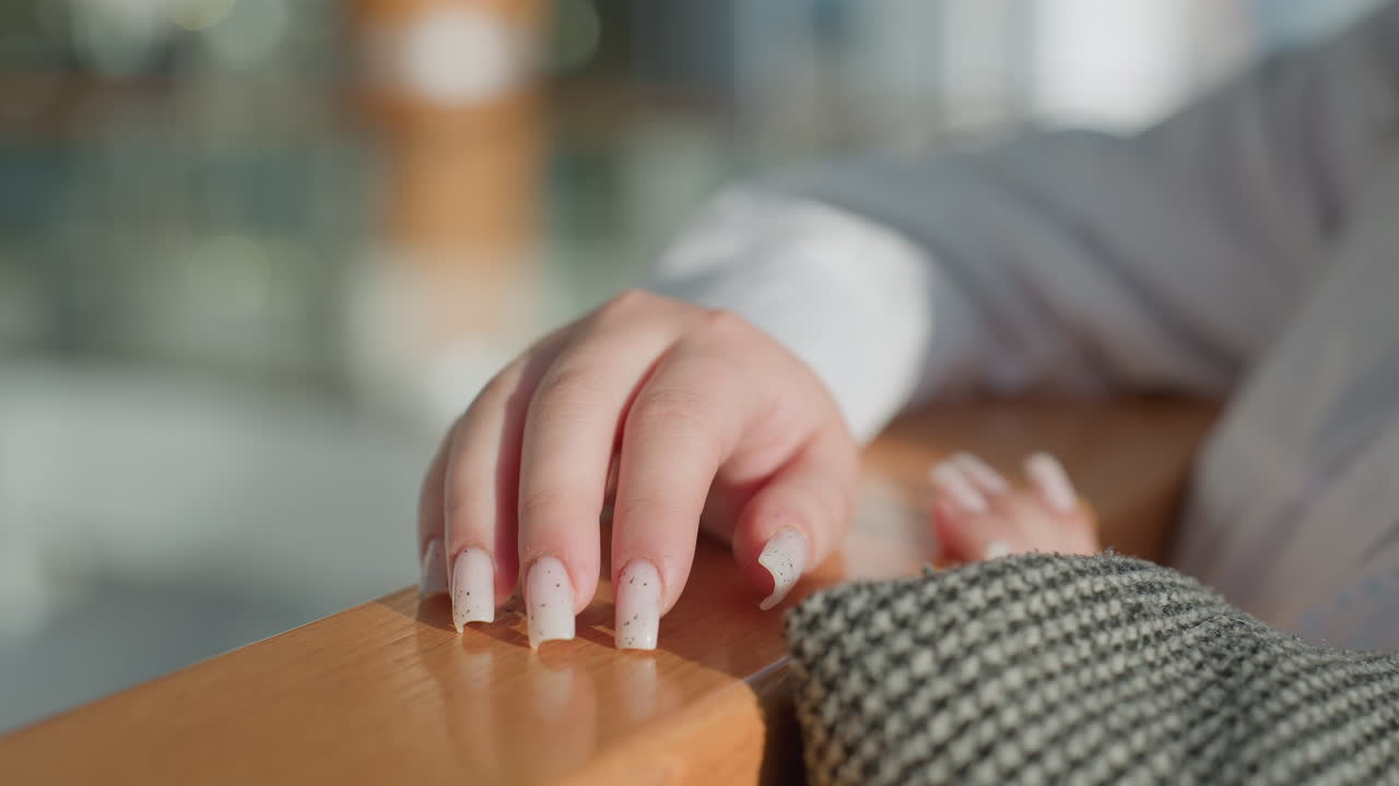 Close up of fair skinned woman gently tapping manicured nails with soft white polish and gold specks on wooden rail in modern shopping mall, with blurred indoor background