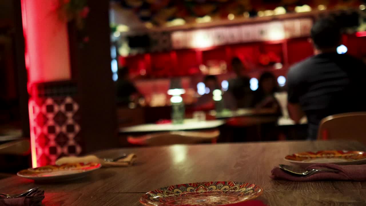 A person stands up and leaves a wooden table set with plates in a warmly lit restaurant with red ambient lighting and festive decor. Camera remains stationary
