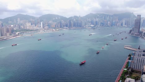 Convoy-of-local-Fishing-boats-causing-in-Hong-Kong-Victoria-bay,-with-city-skyline-in-the-horizon,-Aerial-view