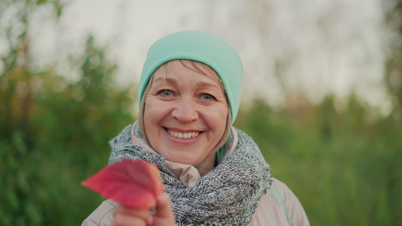 close up of cheerful woman in mint green hat and thick scarf smiling joyfully while admiring autumn leaf in vibrant outdoor setting with soft focus trees and colorful seasonal background