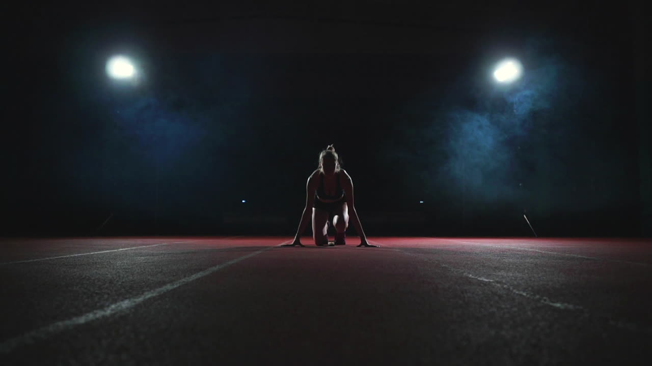 Slender young girl athlete is in position to start running in the pads on the track in slow motion against the smoke