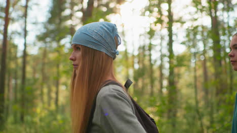 lady wearing blue bandana and gray sweatshirt hikes through sunlit forest, walks thoughtfully, her companion, wearing green shirt, follows close behind, surrounded by greenery and soft sunlight