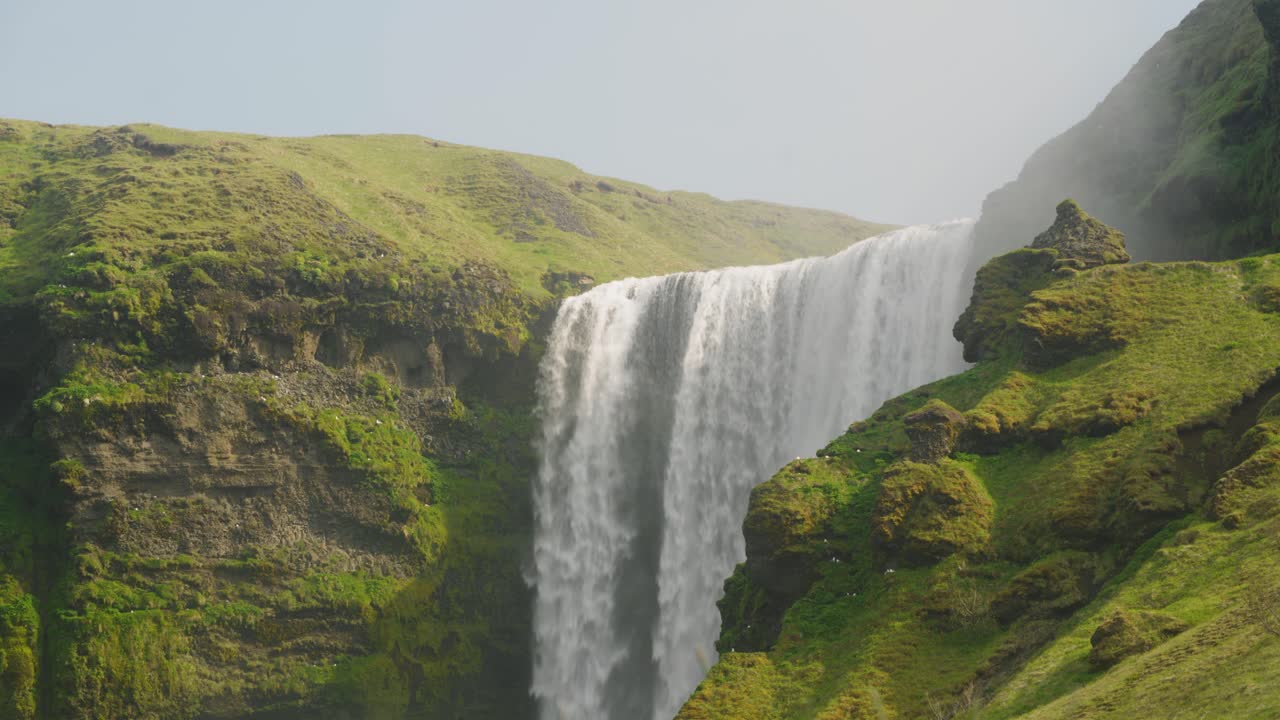 fotografía media de la cascada de skogafoss en islandia, un hermoso día soleado con pájaros volando sobre los acantilados de musgo y las rocas mientras el agua cae