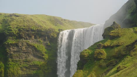 Plano-Medio-De-La-Cascada-De-Skogafoss-En-Islandia,-Hermoso-Día-Soleado-Con-Pájaros-Volando-Sobre-Los-Acantilados-Cubiertos-De-Musgo-Y-Rocas-Mientras-El-Agua-Cae