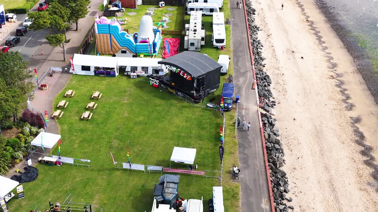 Drone captures fairground rides, tents, and beach along Scottish coast in bright daylight, smooth movement