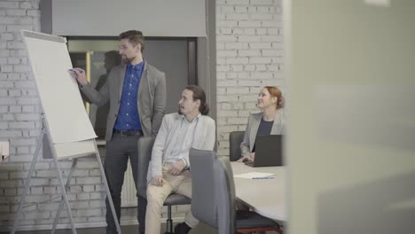 adult caucasian man with grey hair showing graphics on board in open space office to his colleagues. coworkers sitting at the table and discussing project. teamwork, coaching, management, ceo.