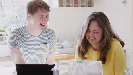 Young-Downs-Syndrome-Couple-Following-Recipe-On-Digital-Tablet-To-Bake-Cake-In-Kitchen-At-Home