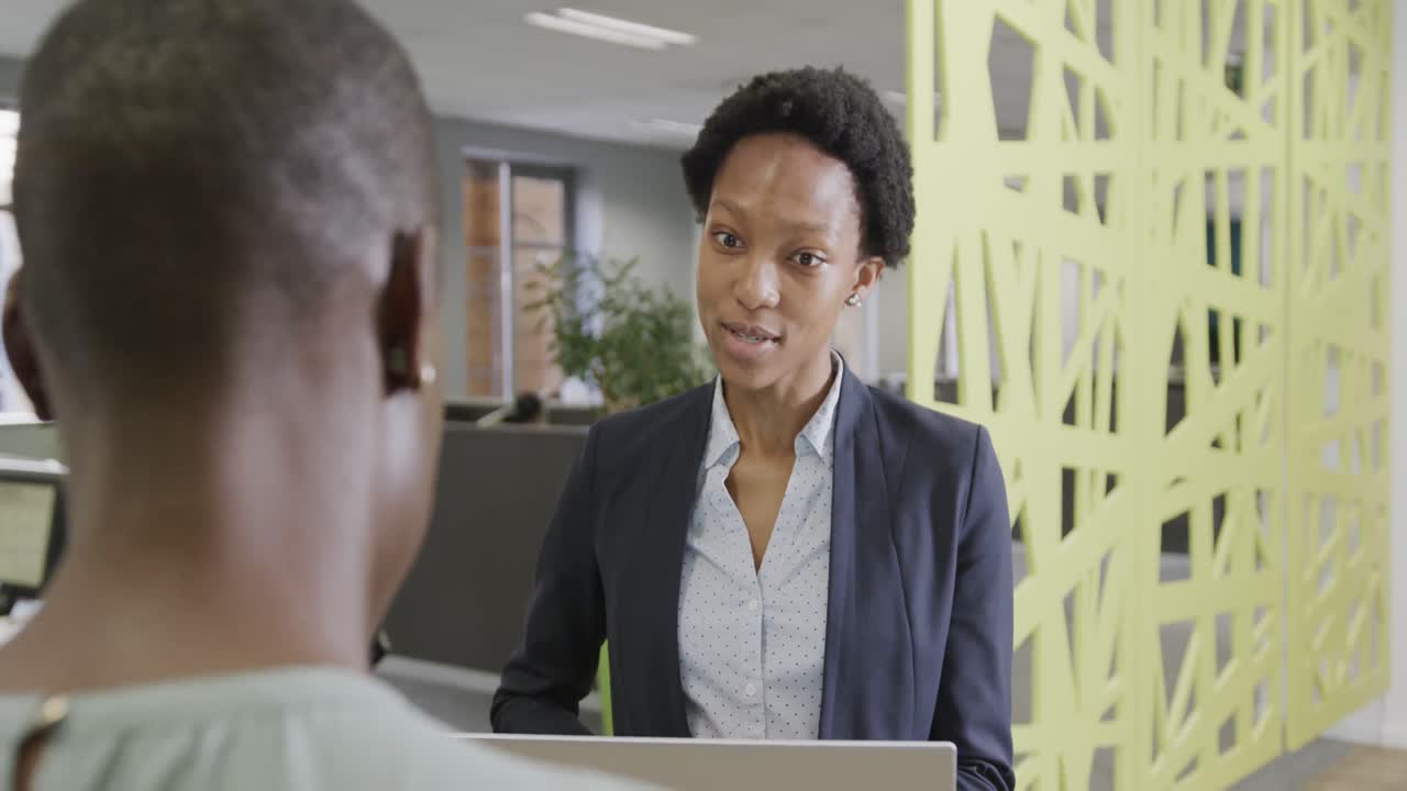Two african american businesswomen discussing work and using laptop in office in slow motion