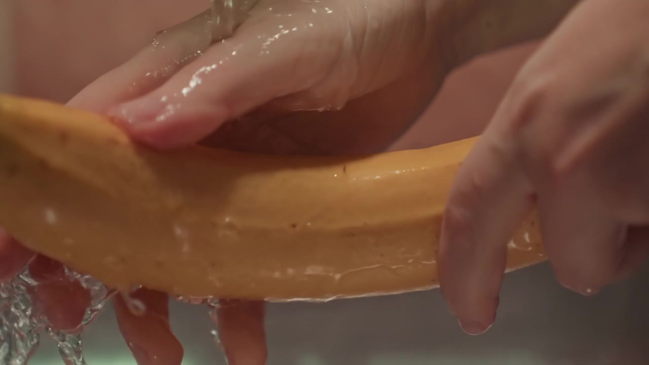 Hands carefully rinse banana under steady tap water while fingers rotate fruit and guide flowing droplets across glossy yellow peel into sink, creating fresh kitchen cleaning scene with visible