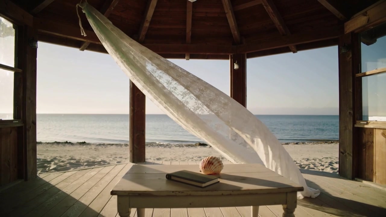Billowing white lace curtain driven by breeze across seaside gazebo, passing closed book and shell
