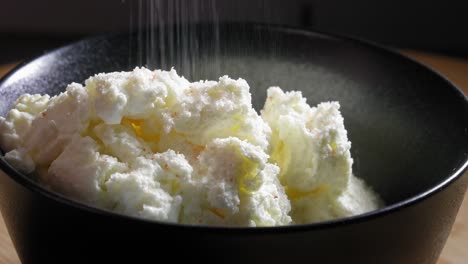 A-close-up-shot-of-a-portion-of-white-low-fat-cheese-in-a-black-ceramic-mixing-bowl,-the-cheese-seasoned-with-a-generous-coating-of-course-salt-and-a-few-millilitres-of-water-is-added-into-the-bowl