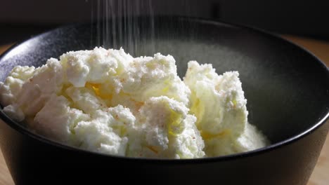 un primer plano de una porción de queso blanco bajo en grasa en un tazón de mezcla de cerámica negra, el queso sazonado con una capa generosa, por supuesto, sal y unos pocos mililitros de agua se agregan al tazón
