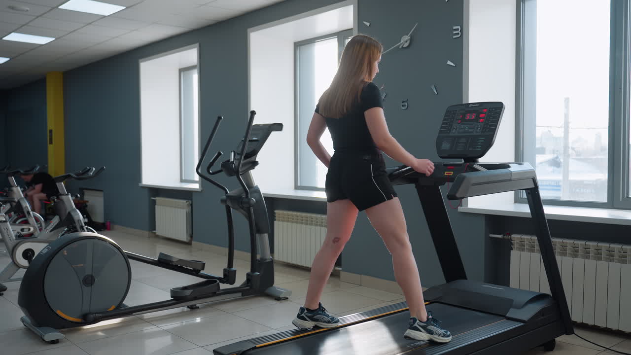 young girl in black gym outfit steps onto treadmill with blinking led display while row of exercise bikes stand in bright modern gym full of natural light and minimalist interior design showing energy