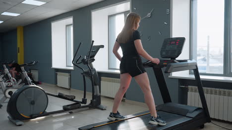 young girl in black gym outfit steps onto treadmill with blinking led display while row of exercise bikes stand in bright modern gym full of natural light and minimalist interior design showing energy