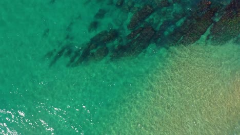 overhead-shot-on-a-sunny-summer-day-of-the-calm-turquoise-sea-in-the-coastal-area-showing-the-orange-sand-and-maroon-rocks-under-the-crystal-clear-water-of-the-coast-of-Cantabria-in-Spain