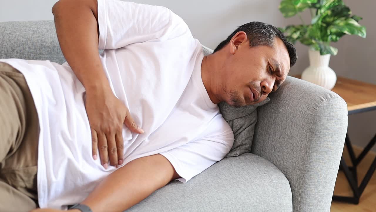 Asian man feeling abdominal pain while lying on a couch indoors
