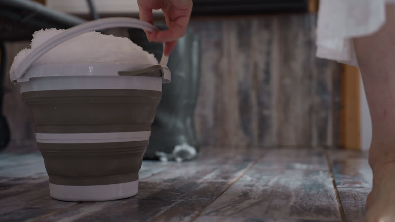 Bare footed lady carrying collapsible bucket heaped with fresh snow across rustic tiled floor, stepping past boots left in corner, droplets glistening under soft winter light
