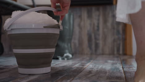 bare footed lady carrying collapsible bucket heaped with fresh snow across rustic tiled floor, stepping past boots left in corner, droplets glistening under soft winter light