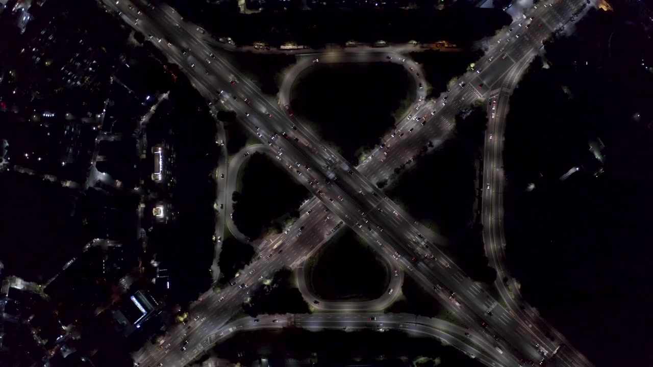Looping ramps and multilane highways form a bright cloverleaf pattern at dark as constant vehicle streams cross in a top-down drone shot. Turcot exchange.