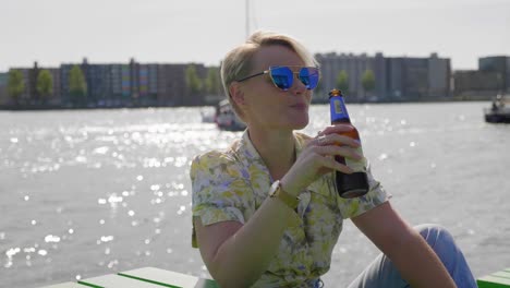 confident young woman drinks a beer in the warm sun, river and city in the background. medium shot.