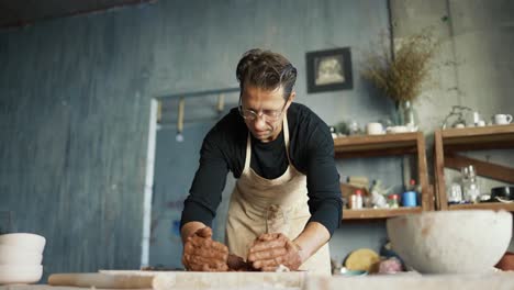 a happy young potter in glasses and and apron is crumpling a piece of clay on a special board in his studio. traditional art