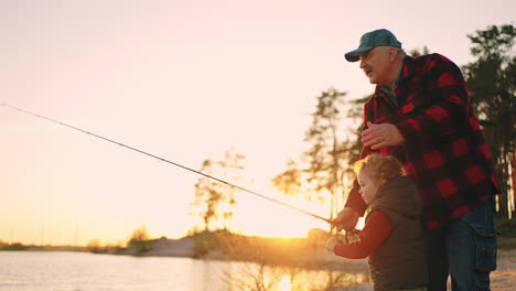 careful grandfather and little grandson are fishing together in river shore in sunset