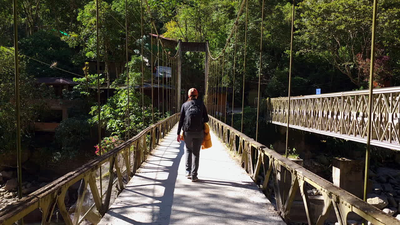 A woman walking on a pedestrian bridge over the Urubamba River. The bridge is part of the scenic route to the ruins of Machu Picchu, a popular travel destination