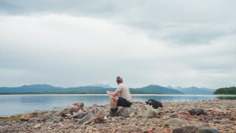 hombre bebiendo una taza de té o café caliente mientras se sienta en una playa rocosa junto al mar con su perro husky siberiano en el parque nacional anderdalen en senja, noruega