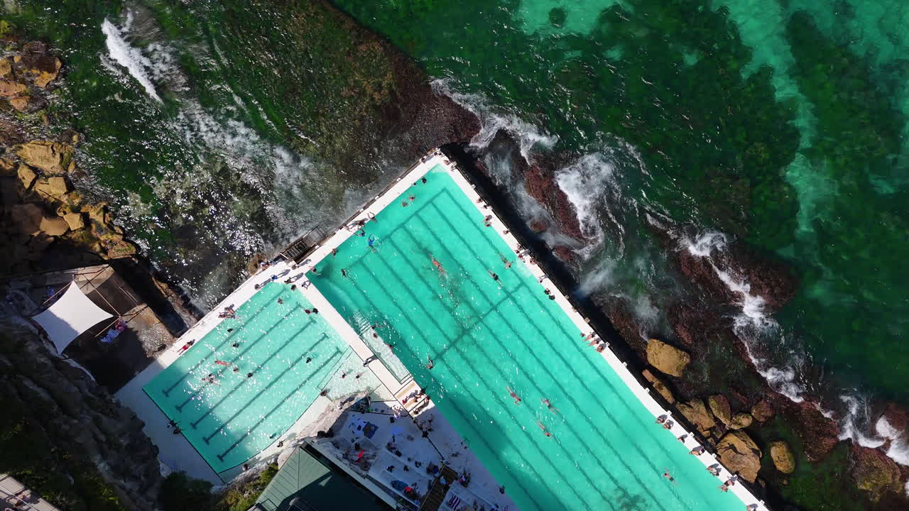 Drone shot of Bondi Beach ocean pool, showcasing people swimming, sunbathing, and relaxing along the coastline. Downward angle drone aerial view, NSW, Australia