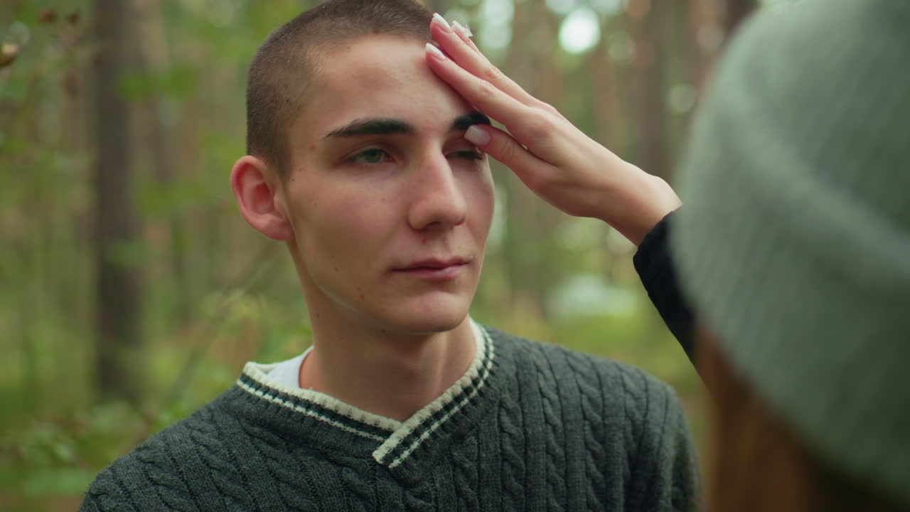 Close up rear view of girl gently removing dirt from boy eye in forest while he playfully covers her face with head warmer, sharing tender moment surrounded by nature and soft green woodland