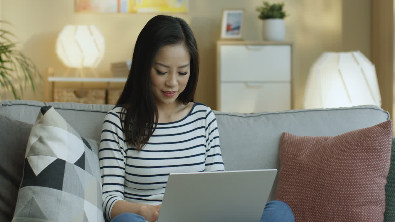 줄무늬 셔츠를 입은 젊은 여성은 랩탑에서 노트북 작업을 하고 거실에 있는 소파에 앉아 생각하고 있습니다 3