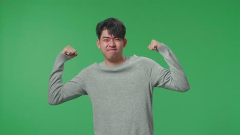 a smiling asian man flex muscle while standing in front of green screen background