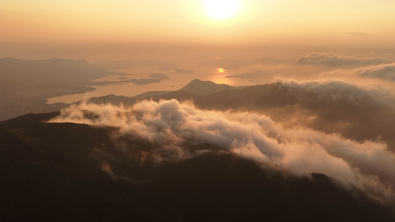 Aerial view of sunrise over misty mountains and islands in Hong Kong, golden sunlight reflecting on the sea, dramatic clouds flowing across forested peaks in serene landscape