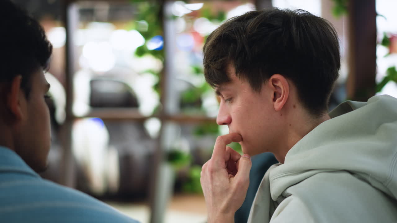 Side view of white boy touching cheek while looking intently in one direction where phone lies with blurred people in background, quiet focus and subtle emotion captured in casual setting