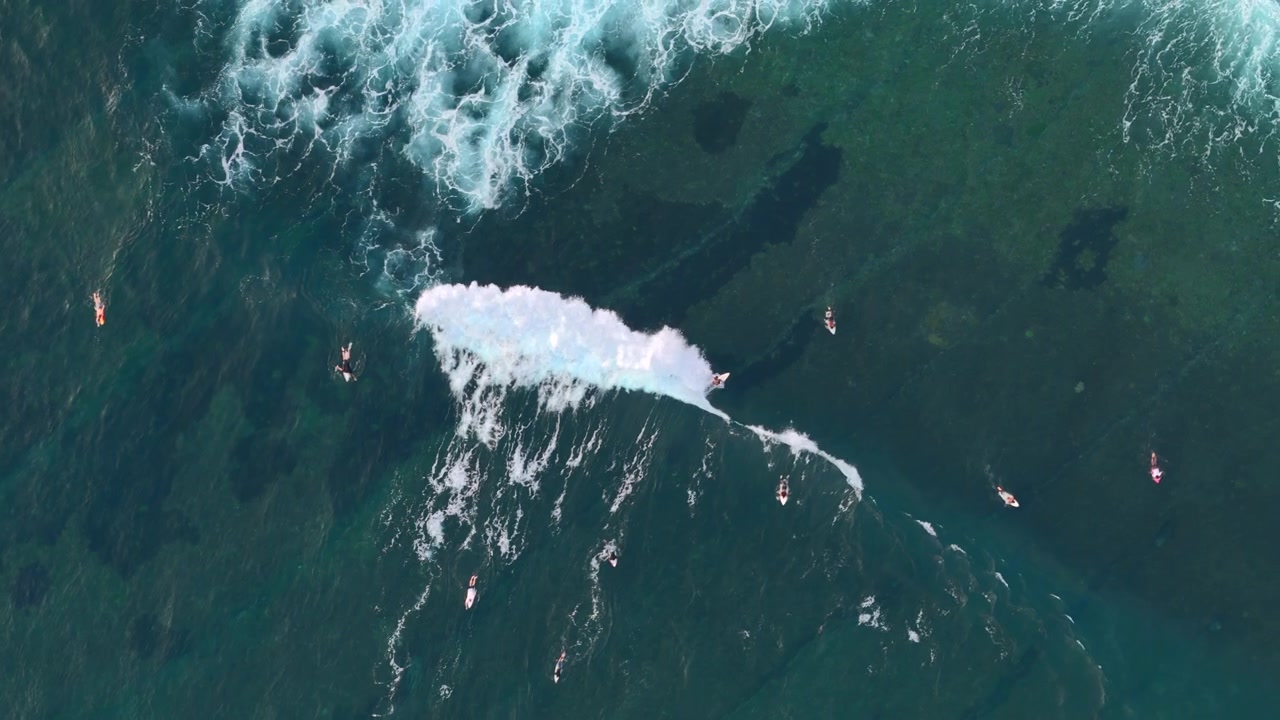 Vista aerea dall'alto di surfisti che aspettano e cavalcano le onde sopra l'acqua scura della barriera corallina. Scena oceanica tropicale con schiuma bianca, texture del mare limpida, viaggi d'avventura e stile di vita costiero attivo.